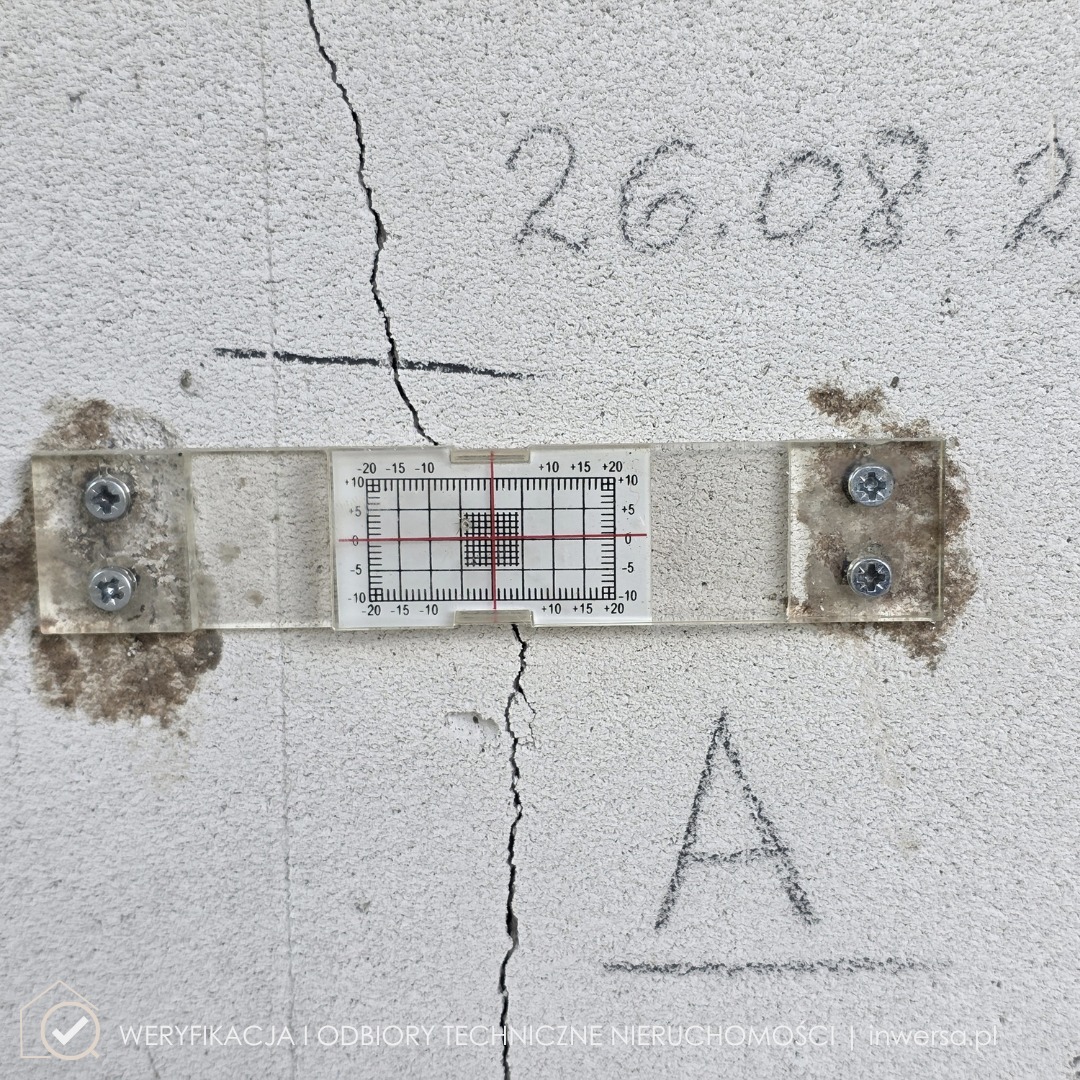 Pęknięcie ściany z zamontowanym szczelinomierzem, data pomiaru ołówkiem. Narzędzie do pomiaru rozwarcia rys, analiza stanu technicznego budynku.