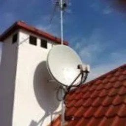 Antena satelitarna zamontowana na dachu z czerwonej dachówki obok białego komina, widoczna również antena naziemna na maszcie, błękitne niebo w tle.
