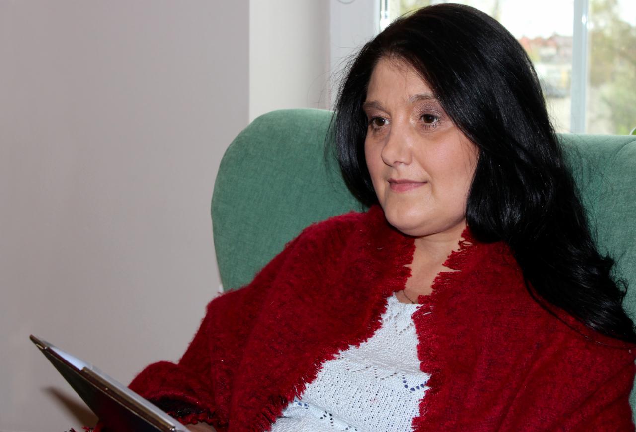 Kobieta siedząca w zielonym fotelu, ubrana w czerwony szal, trzymająca tablet w dłoni, zamyślony wyraz twarzy, widok z boku.