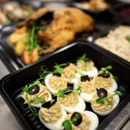 Gotowe dania w czarnych pojemnikach cateringowych: faszerowane jajka z oliwkami i kiełkami, pieczona kaczka z jabłkami, sałatka jarzynowa.