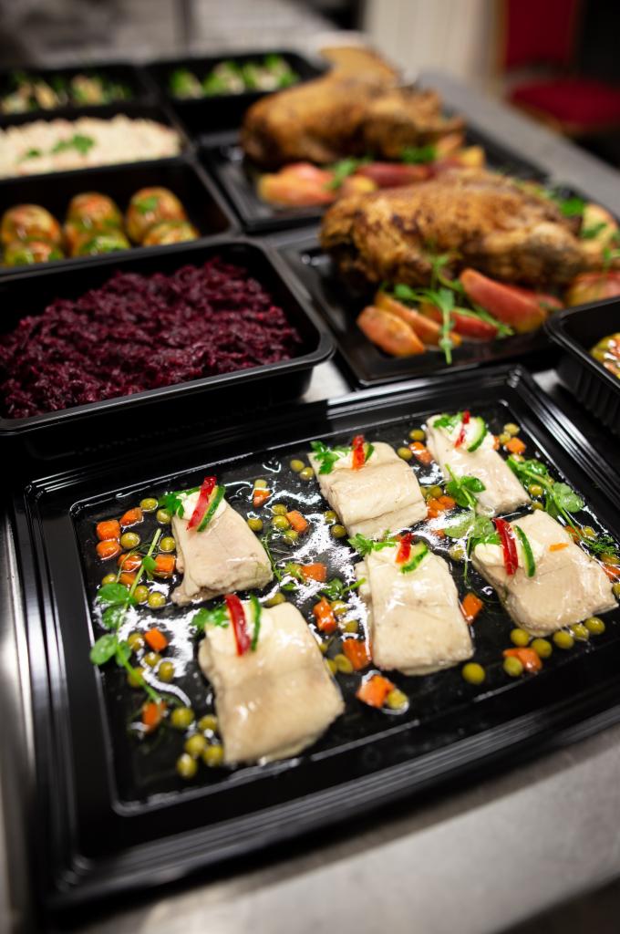 Gotowe dania w czarnych pojemnikach cateringowych: śledź w galarecie z warzywami, pieczona ryba z jabłkami i sałatą, buraczki, ziemniaki i ryż.