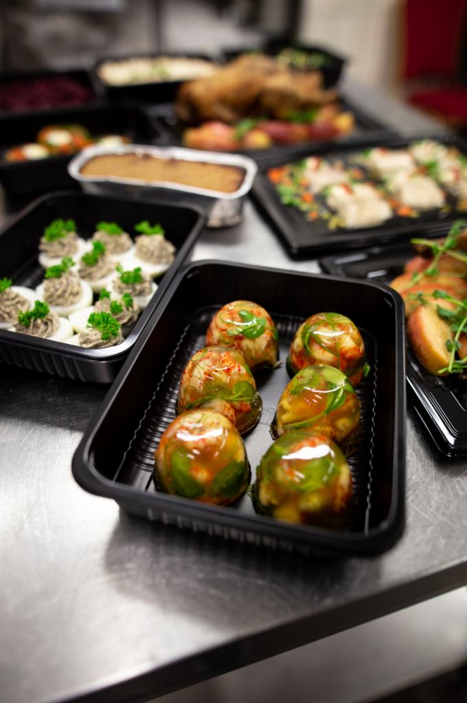 Przygotowane do transportu galaretki mięsne w czarnych pojemnikach, obok jajka faszerowane i pieczone mięso, widoczne na metalowym stole.