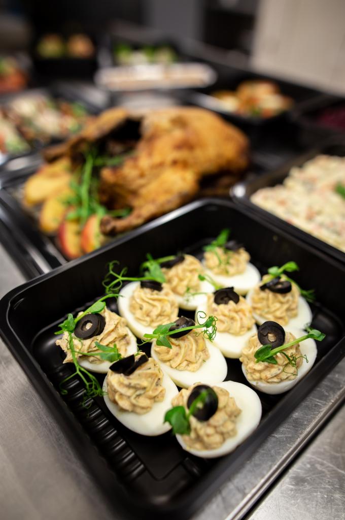 Gotowe dania w czarnych pojemnikach cateringowych: faszerowane jajka z oliwkami i kiełkami, pieczona kaczka z jabłkami, sałatka jarzynowa.