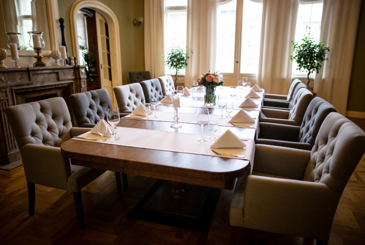 Uroczysta aranżacja stołu z szarymi krzesłami z pikowaniem, białymi serwetkami, kieliszkami i kwiatami, ustawiona w eleganckim wnętrzu z kominkiem i dużymi oknami.