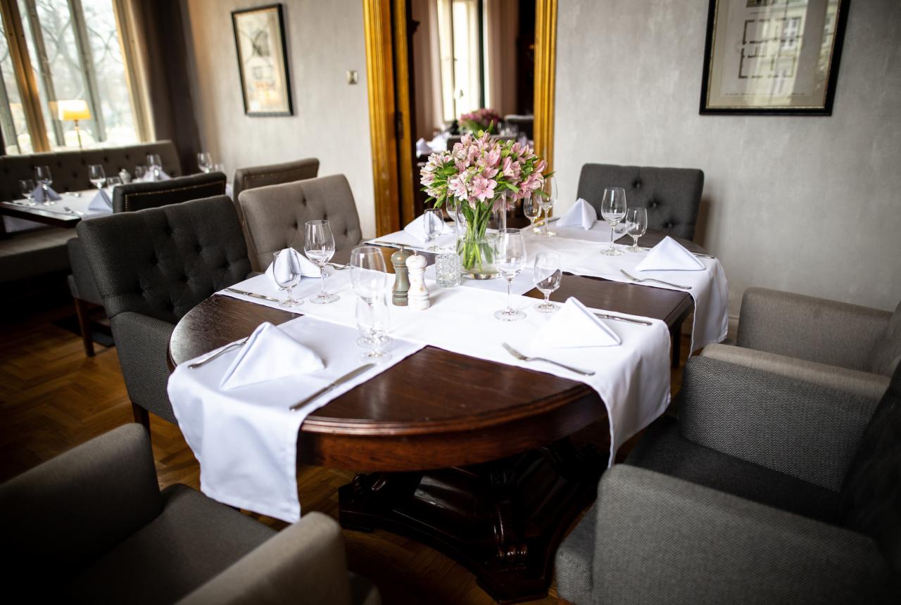 Elegancko nakryty stół z ciemnego drewna w restauracji, białe obrusy i serwetki, szklane kieliszki, wazon z różowymi kwiatami, otoczony szarymi fotelami.