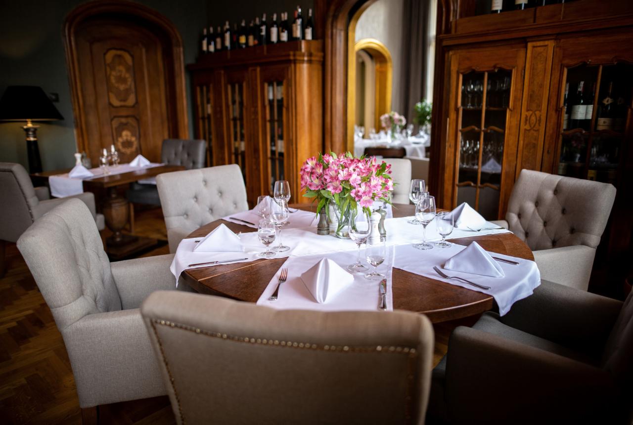 Elegancko nakryty okrągły stół z bukietem różowych kwiatów w szklanym wazonie, otoczony tapicerowanymi krzesłami w restauracji z drewnianymi kredensami w tle.