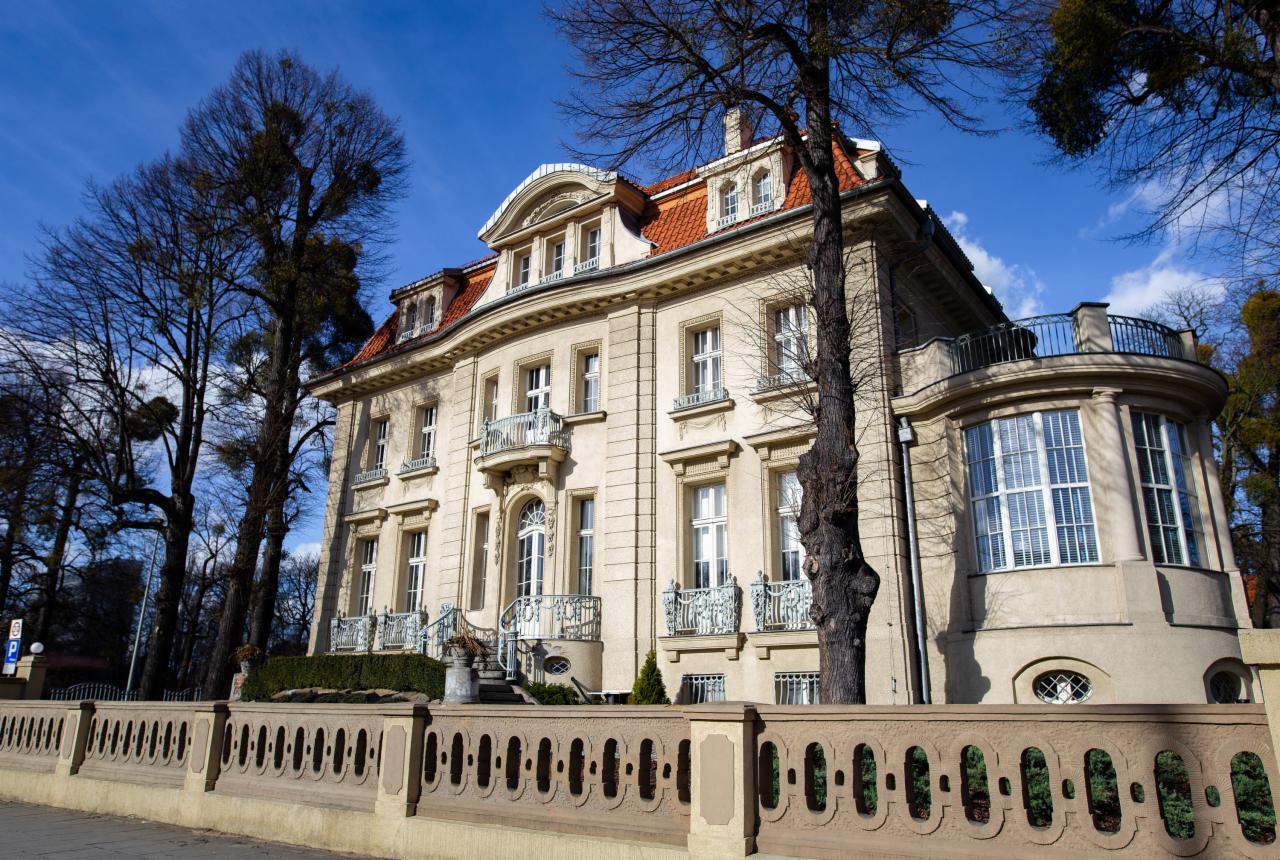 Elegancka, trzypiętrowa willa w stylu klasycystycznym z czerwonym dachem, balkonami z kutymi balustradami i ozdobnym ogrodzeniem z betonu, otoczona drzewami bez liści pod błękitnym niebem.