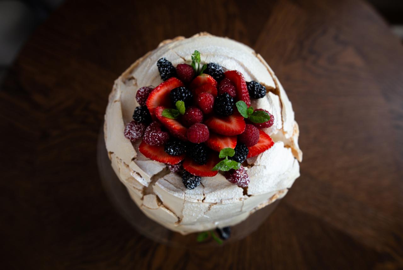 Beza Pavlova z truskawkami, malinami, jeżynami i listkami mięty, posypana cukrem pudrem, na drewnianym stole, widok z góry.