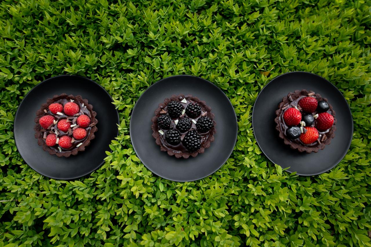 Trzy czekoladowe tartaletki na czarnych talerzach, każda z innym rodzajem owoców: malinami, jeżynami i mieszanką truskawek z borówkami, ułożone na tle zielonych liści.