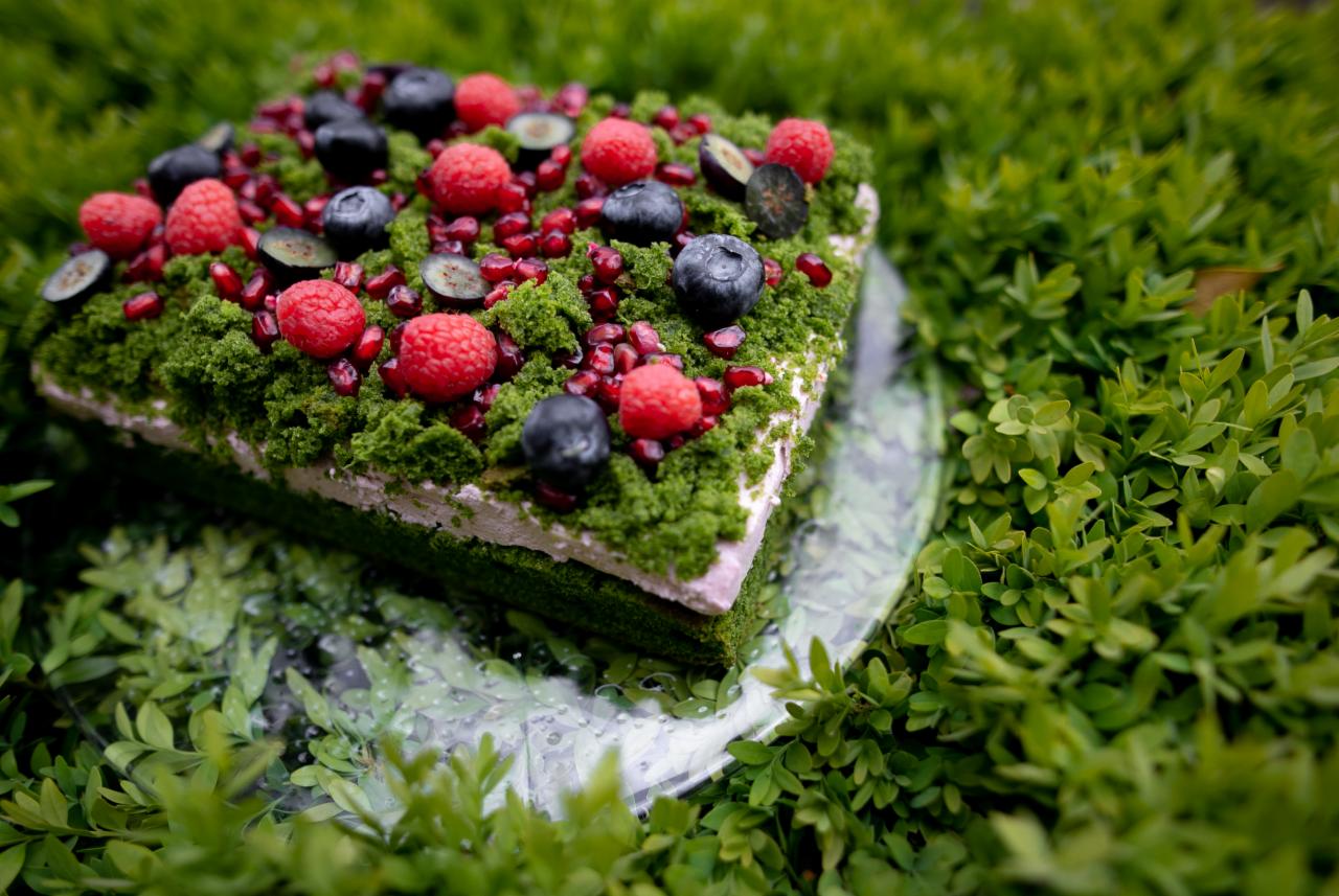 Kwadratowy tort z zielonym kremem przypominającym mech, udekorowany malinami, borówkami i granatem, umieszczony na szklanej paterze otoczonej zielenią.