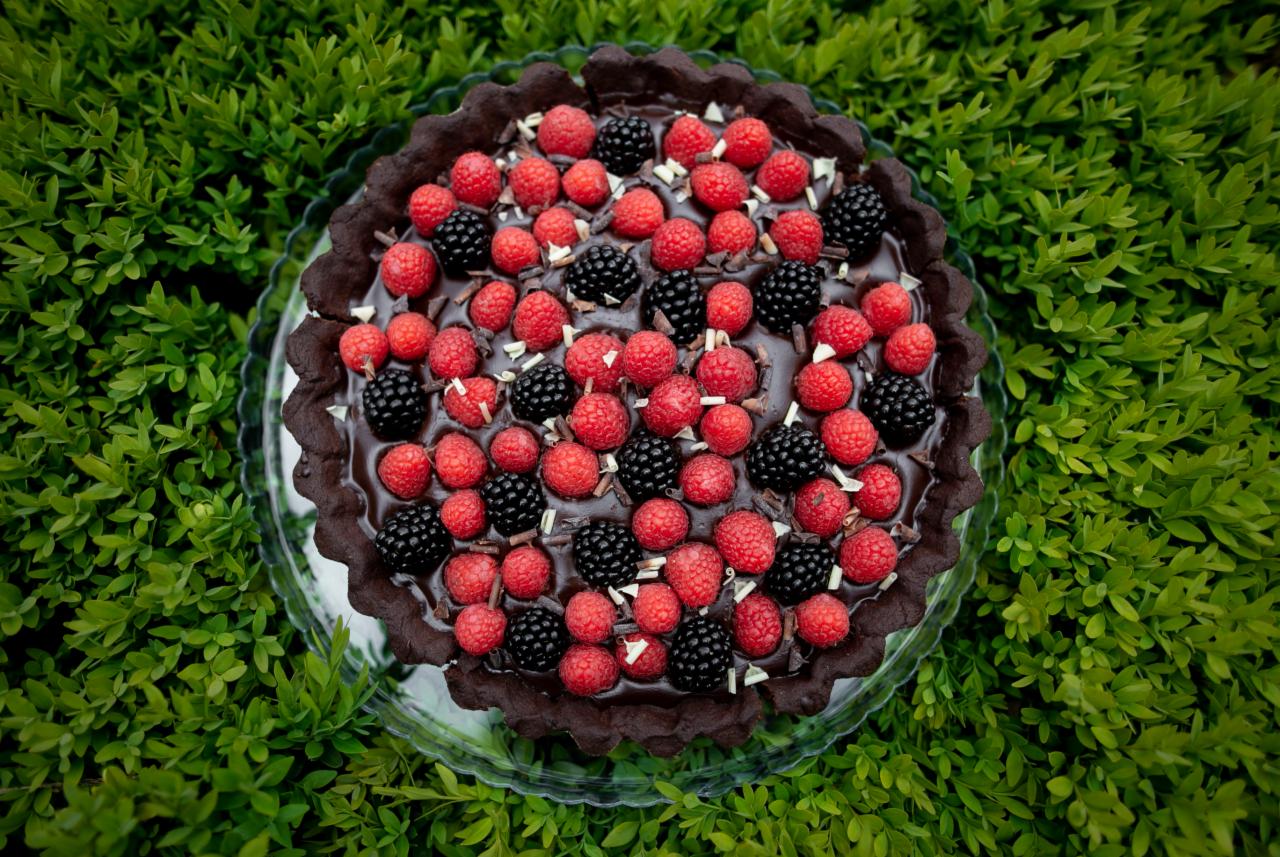 Czekoladowa tarta z malinami i jeżynami na szklanym talerzu, umieszczona na tle zielonego krzewu.