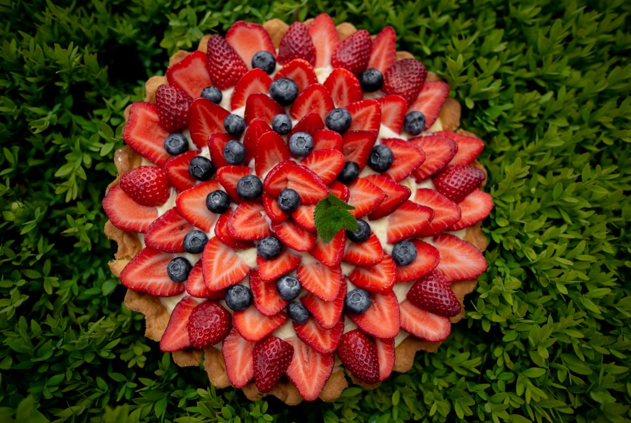 Tarta z kremem, udekorowana świeżymi truskawkami i borówkami, z pojedynczym listkiem mięty na wierzchu, na tle zielonych krzewów.