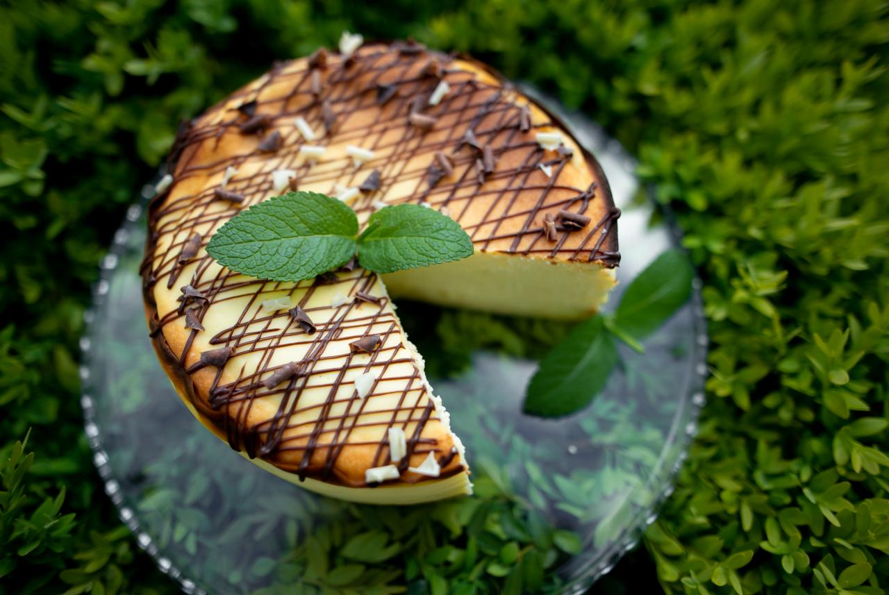 Serowy tort udekorowany czekoladą i miętą, wyeksponowany na szklanym talerzu na tle zielonych krzewów.