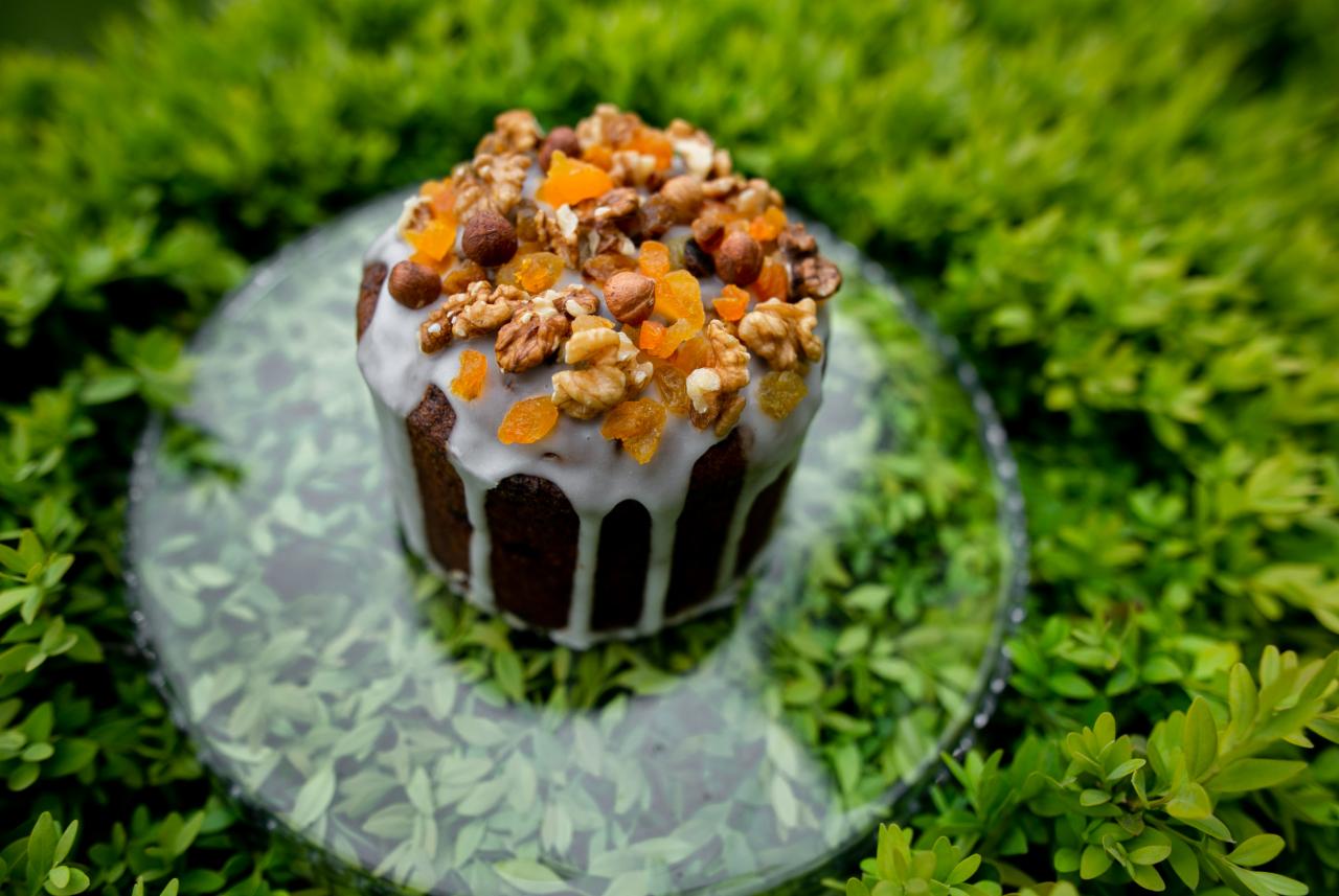 Babka wielkanocna polana lukrem i posypana orzechami włoskimi, laskowymi i kandyzowaną skórką pomarańczową, na szklanym talerzu, w tle zielona roślinność.