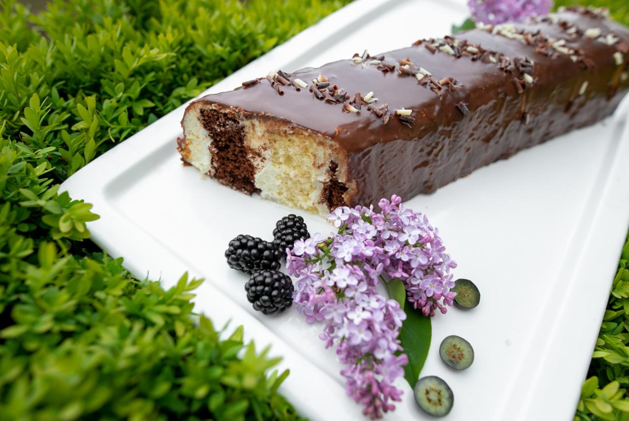 Przekrojone ciasto zebra polane czekoladą, posypane wiórkami, udekorowane jeżynami, gałązką bzu i przekrojonymi borówkami na białej tacy, z zielonym tłem.
