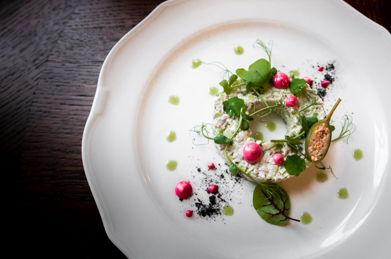 Wyrafinowana kompozycja sałatki w kształcie pierścienia, udekorowana różowymi kulkami, zieleniną i kaparem na białym talerzu z delikatnymi kroplami sosu.