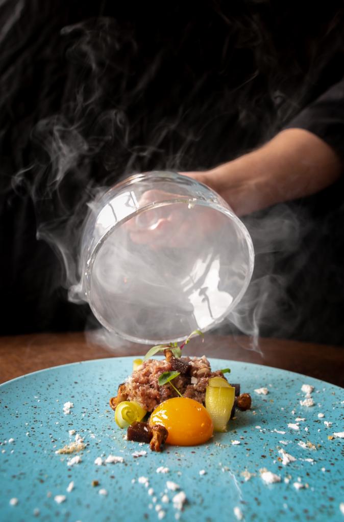Wykwintne danie z tatarem, żółtkiem i marynowanymi warzywami na turkusowym talerzu, unoszący się dym spod szklanego klosza trzymanego przez kucharza.