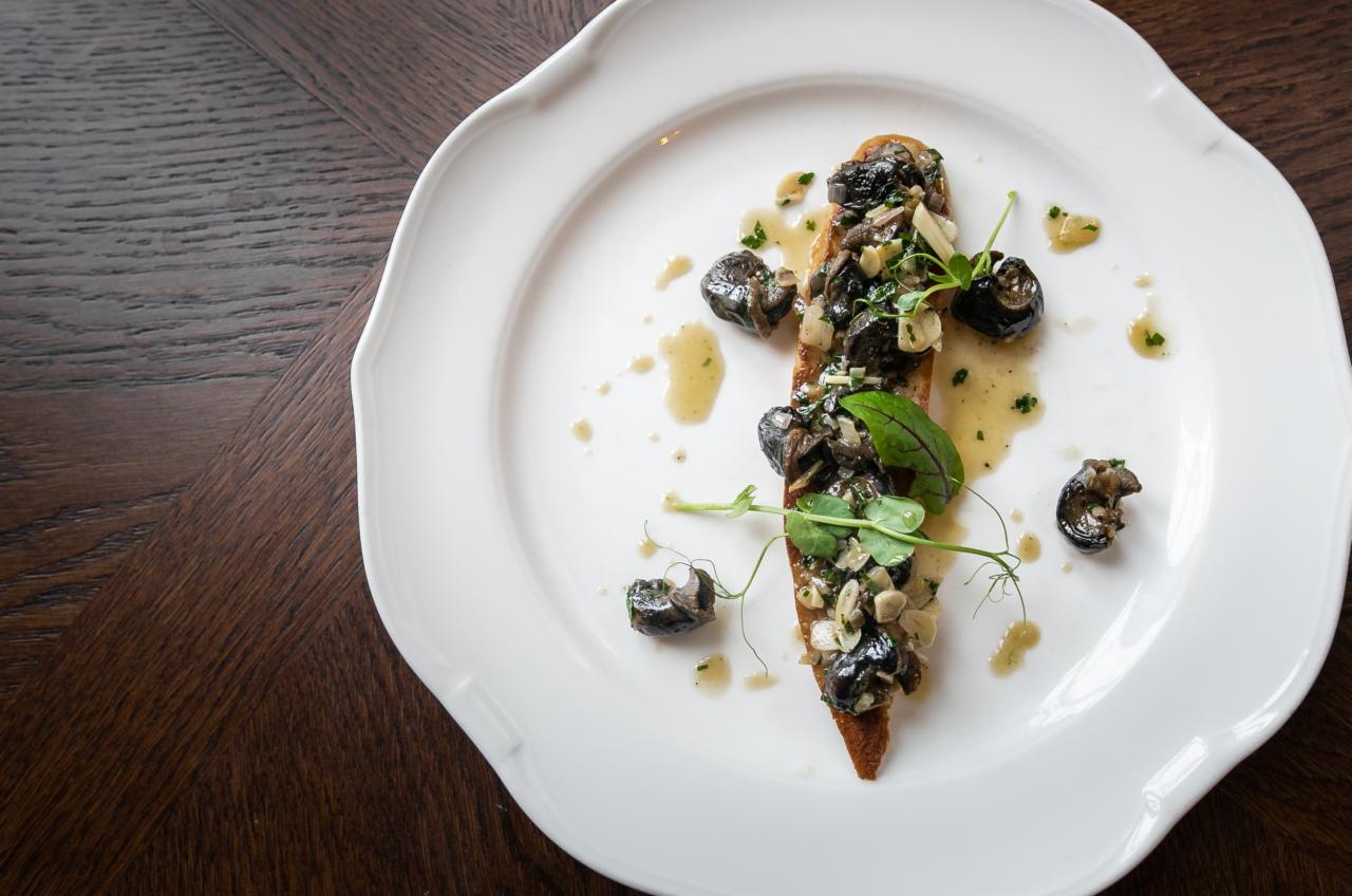 Elegancko podane grzanki z ciemnymi ślimakami, ziołami i sosem na białym talerzu z falistym brzegiem, umieszczonym na ciemnym, drewnianym stole.