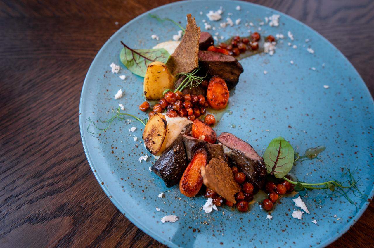 Elegancko podane danie mięsne z pieczonymi warzywami i dekoracjami na turkusowym talerzu, widok z góry, drewniany stół w tle.