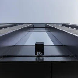 Kamera monitoringu zamontowana na elewacji budynku, widok z dołu na tle okna i nieba.