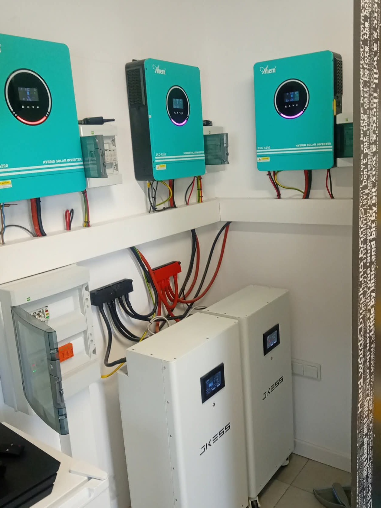 Kompletna instalacja fotowoltaiczna w pomieszczeniu: turkusowe inwertery hybrydowe, białe akumulatory JKESS, rozdzielnica elektryczna. Widoczne okablowanie i elementy montażowe.