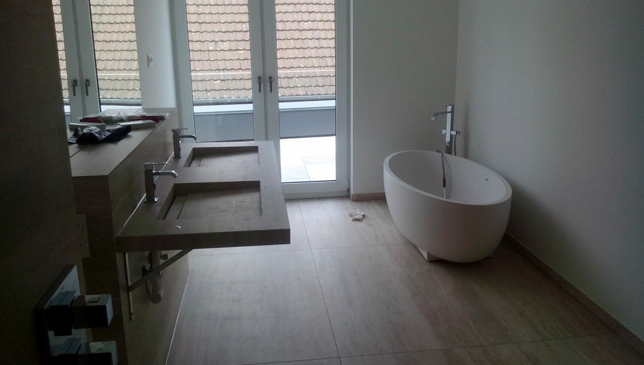 Nowoczesna łazienka w trakcie remontu z podwójną umywalką z szarego kamienia, wolnostojącą białą wanną i beżowymi płytkami podłogowymi, widoczne narzędzia na blacie umywalki.