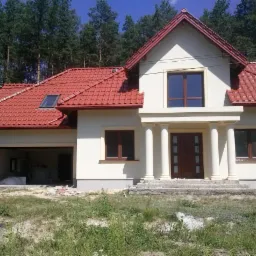 Nowo wybudowany dom jednorodzinny z czerwoną dachówką i kremową elewacją, widoczne kolumny przy wejściu i częściowo otwarty garaż z materiałami budowlanymi w środku.