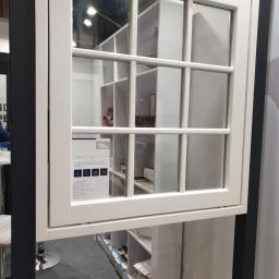 Eko-Okna Scandinavia Sp. z.o.o - Białe okno z podziałami szprosowymi, prezentowane na ekspozycji, z widoczną naklejką informacyjną producenta. W tle regały z ekspozycją produktów.