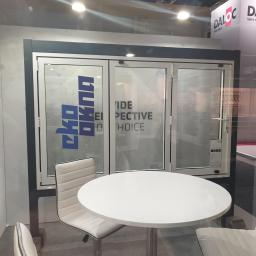 Eko-Okna Scandinavia Sp. z.o.o - Trzyczęściowe białe drzwi balkonowe z widocznym logo producenta i hasłem reklamowym, ustawione na ekspozycji targowej z okrągłym białym stolikiem i krzesłami w stylu skandynawskim.