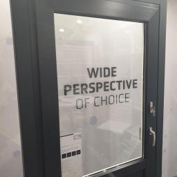 Eko-Okna Scandinavia Sp. z.o.o - Szare drzwi zewnętrzne z szybą z naklejonym napisem 'WIDE PERSPECTIVE OF CHOICE' oraz kluczami w zamku.