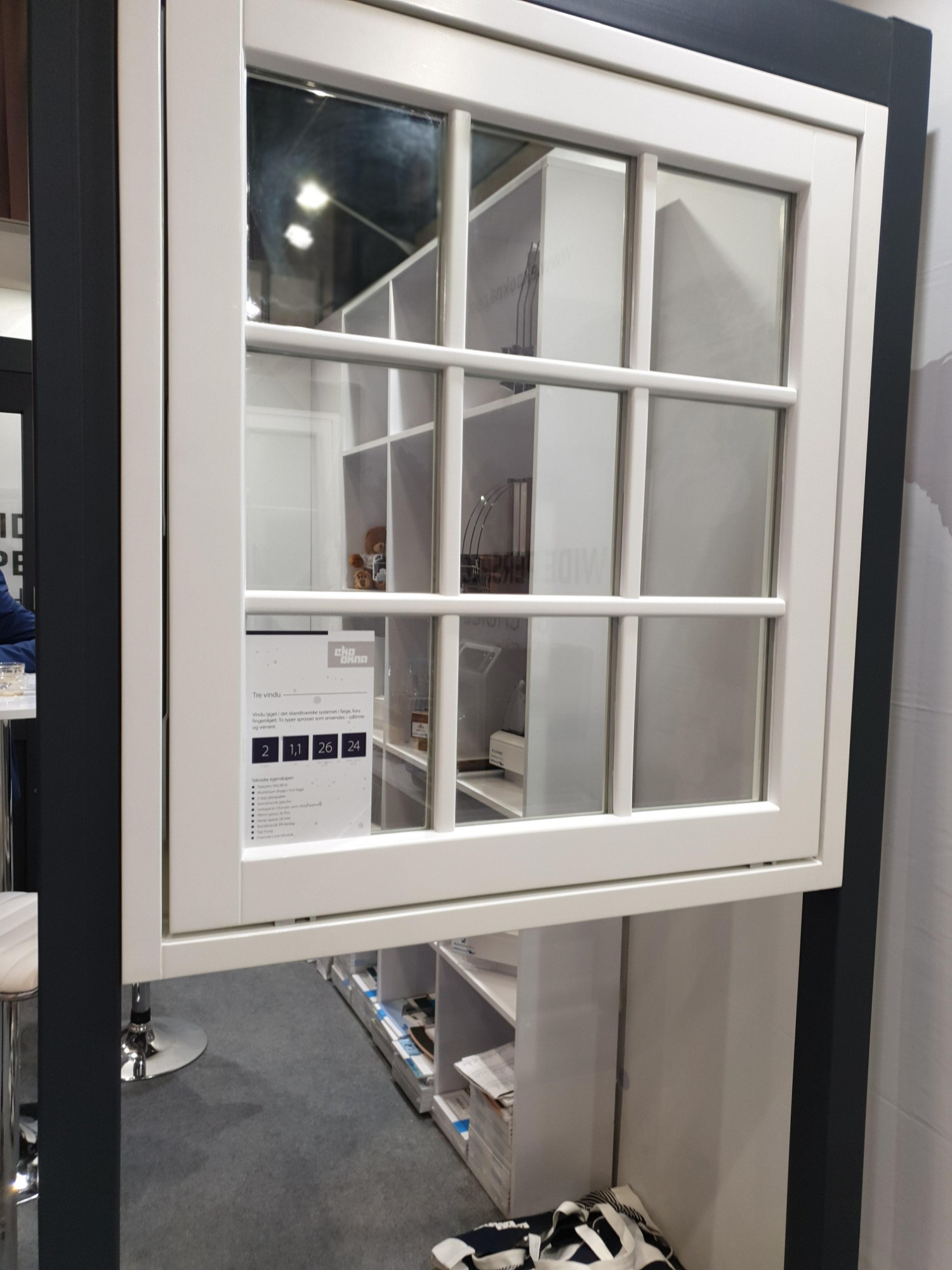 Białe okno z podziałami szprosowymi, prezentowane na ekspozycji, z widoczną naklejką informacyjną producenta. W tle regały z ekspozycją produktów.