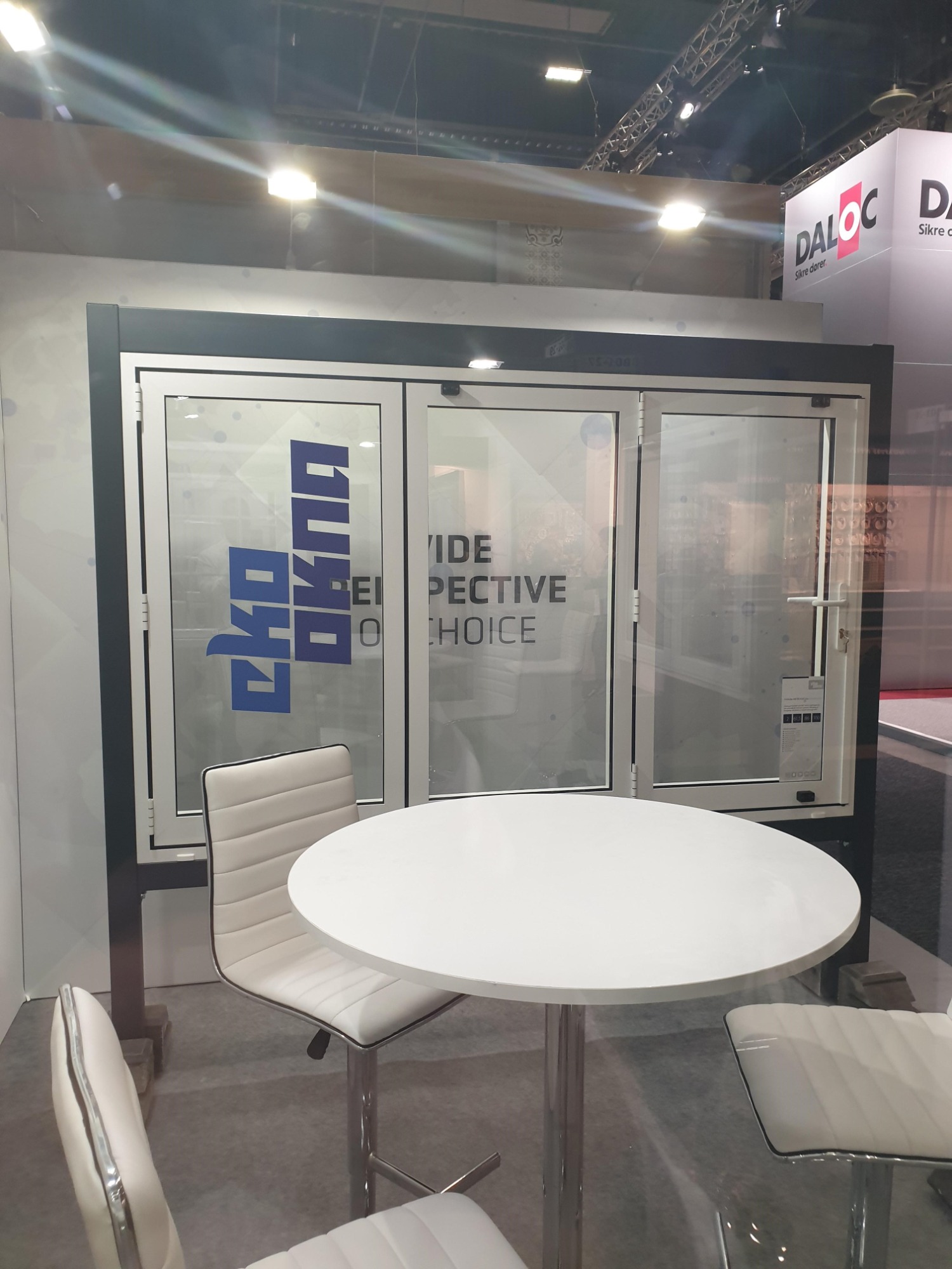 Trzyczęściowe białe drzwi balkonowe z widocznym logo producenta i hasłem reklamowym, ustawione na ekspozycji targowej z okrągłym białym stolikiem i krzesłami w stylu skandynawskim.