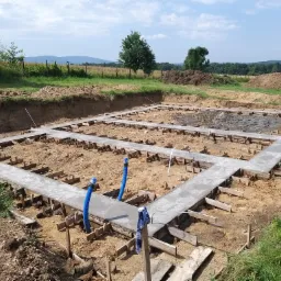 Wylane betonowe ławy fundamentowe w szalunku z deskami, widoczne niebieskie rury instalacyjne wystające z betonu, teren przygotowany pod budowę domu z widokiem na pagórkowaty krajobraz w tle.