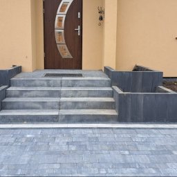 Ma-Pa Projekt - Schody zewnętrzne z szarej kostki brukowej prowadzące do brązowych drzwi wejściowych, z donicami po bokach i fragmentem ułożonej nawierzchni z kostki.