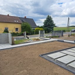 Ma-Pa Projekt - W trakcie układania ścieżki z płyt chodnikowych w ogrodzie, widoczny pracownik z taczką, przygotowane podłoże i częściowo ułożone płyty. W tle dom jednorodzinny i brama wjazdowa.