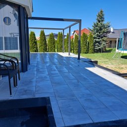 Ma-Pa Projekt - Nowoczesny taras z szarych płyt, z metalową pergolą w tle, widok na ogród z tujami i trampoliną.