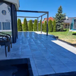 Nowoczesny taras z szarych płyt, z metalową pergolą w tle, widok na ogród z tujami i trampoliną.