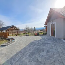 Ułożona szara kostka brukowa przed domem z drewnianą pergolą w tle, widoczny mini-koparka i góry w oddali.