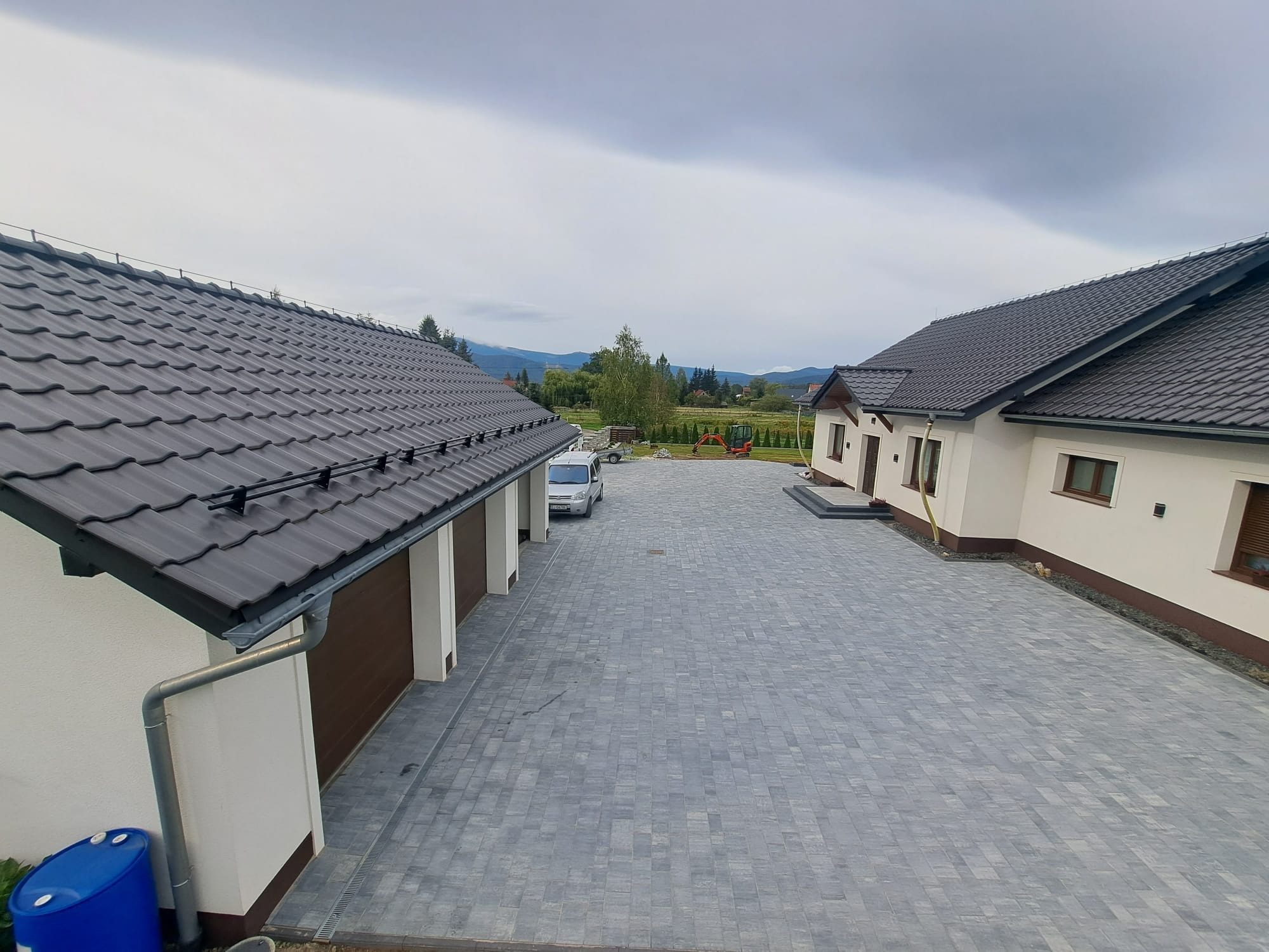 Widok z góry na posesję z szarą kostką brukową, dwoma budynkami z ciemnymi dachami i widokiem na góry w tle, w pochmurny dzień.