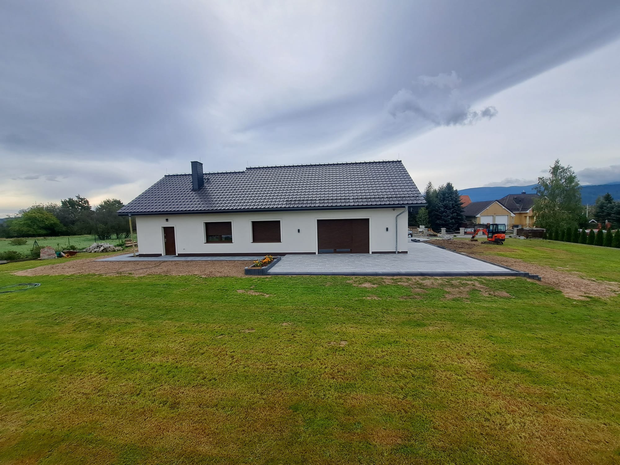 Nowoczesny dom z szarą kostką brukową na podjeździe, widoczny mały pomarańczowy traktor w tle, trawnik z miejscami lekko przesuszony, pochmurne niebo.