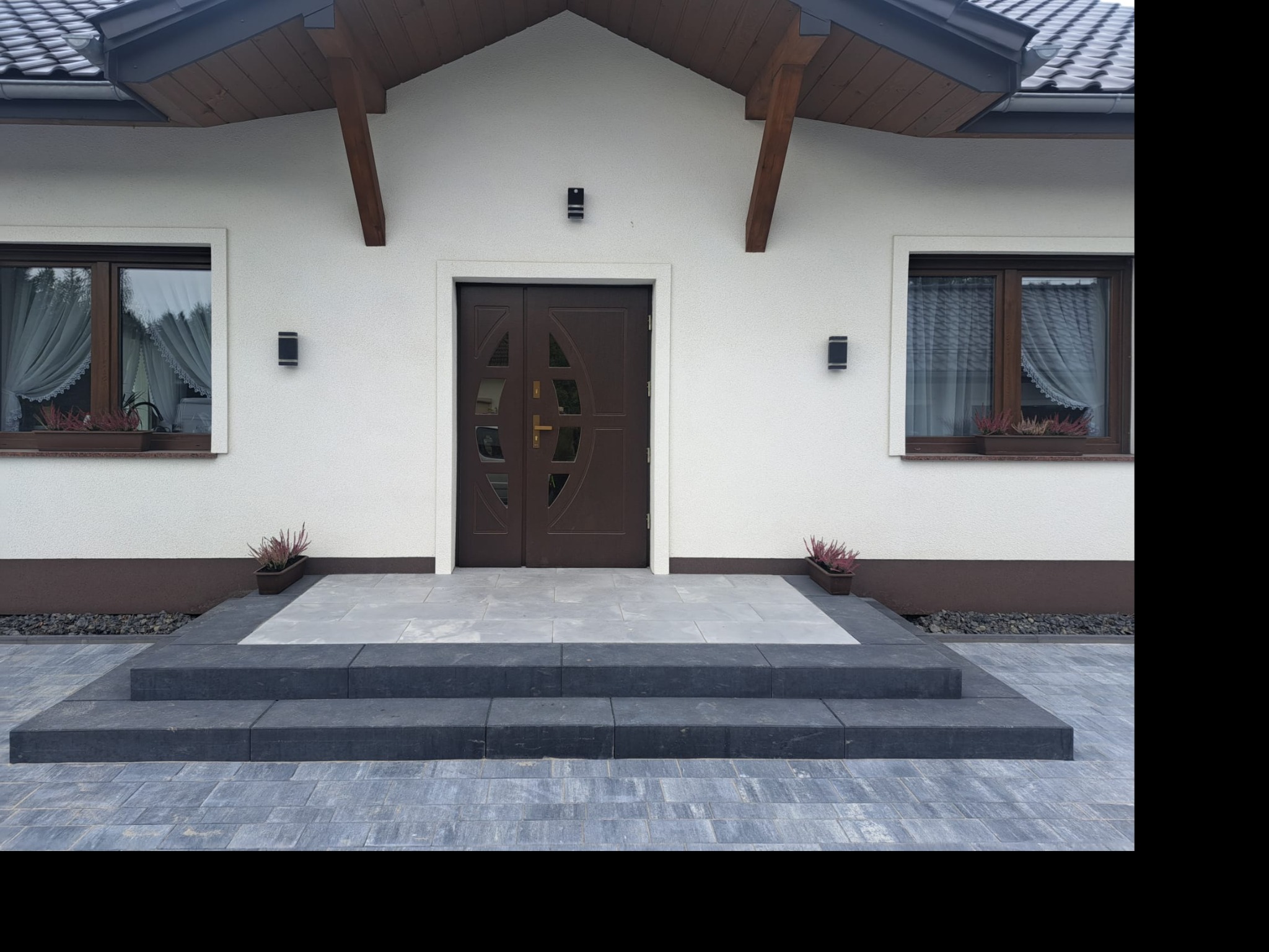 Nowoczesne schody wejściowe z ciemnoszarej kostki brukowej prowadzące do brązowych drzwi wejściowych domu z białymi ścianami, ozdobione doniczkami z wrzosami w oknach.