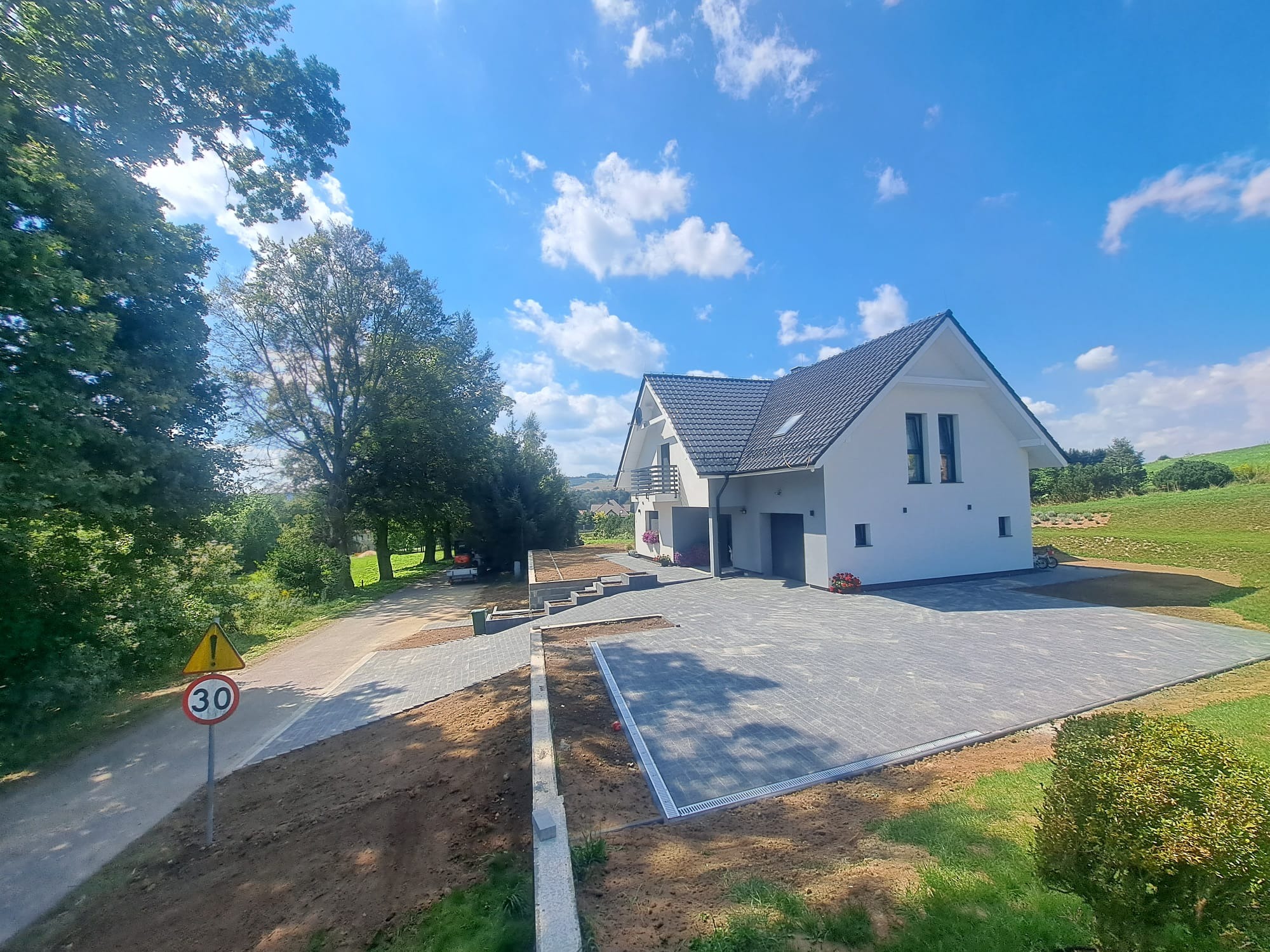 Nowoczesny dom z szarej kostki brukowej na podjeździe i ścieżce, widok z góry, znak drogowy '30' w tle, błękitne niebo z chmurami.