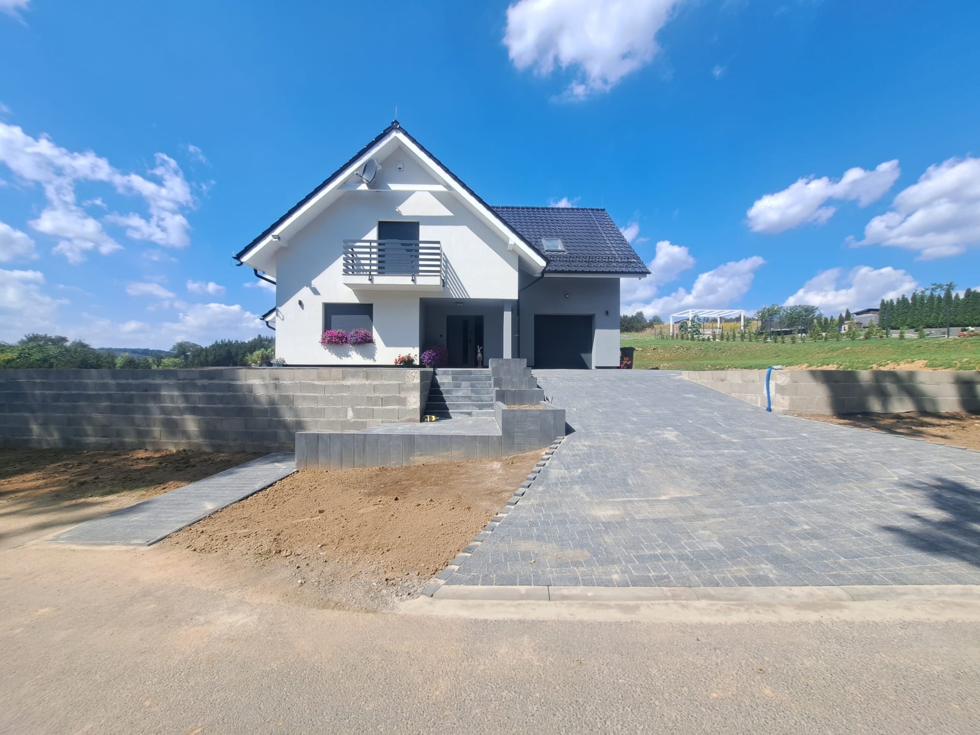 Nowoczesny dom z podjazdem wyłożonym szarą kostką brukową, schody wejściowe z tego samego materiału, mur oporowy z betonowych bloczków, trawnik w tle, błękitne niebo z chmurami.