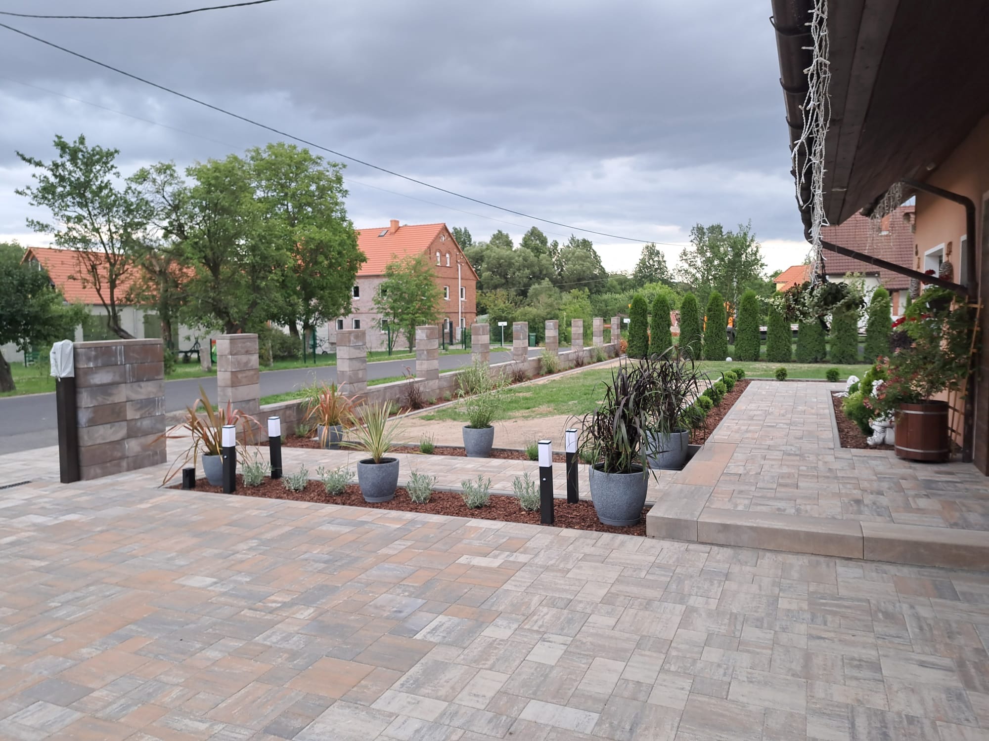 Ułożona kostka brukowa w odcieniach szarości i beżu, z oświetleniem ogrodowym i roślinami w donicach, tworząca estetyczny podjazd i ścieżkę.