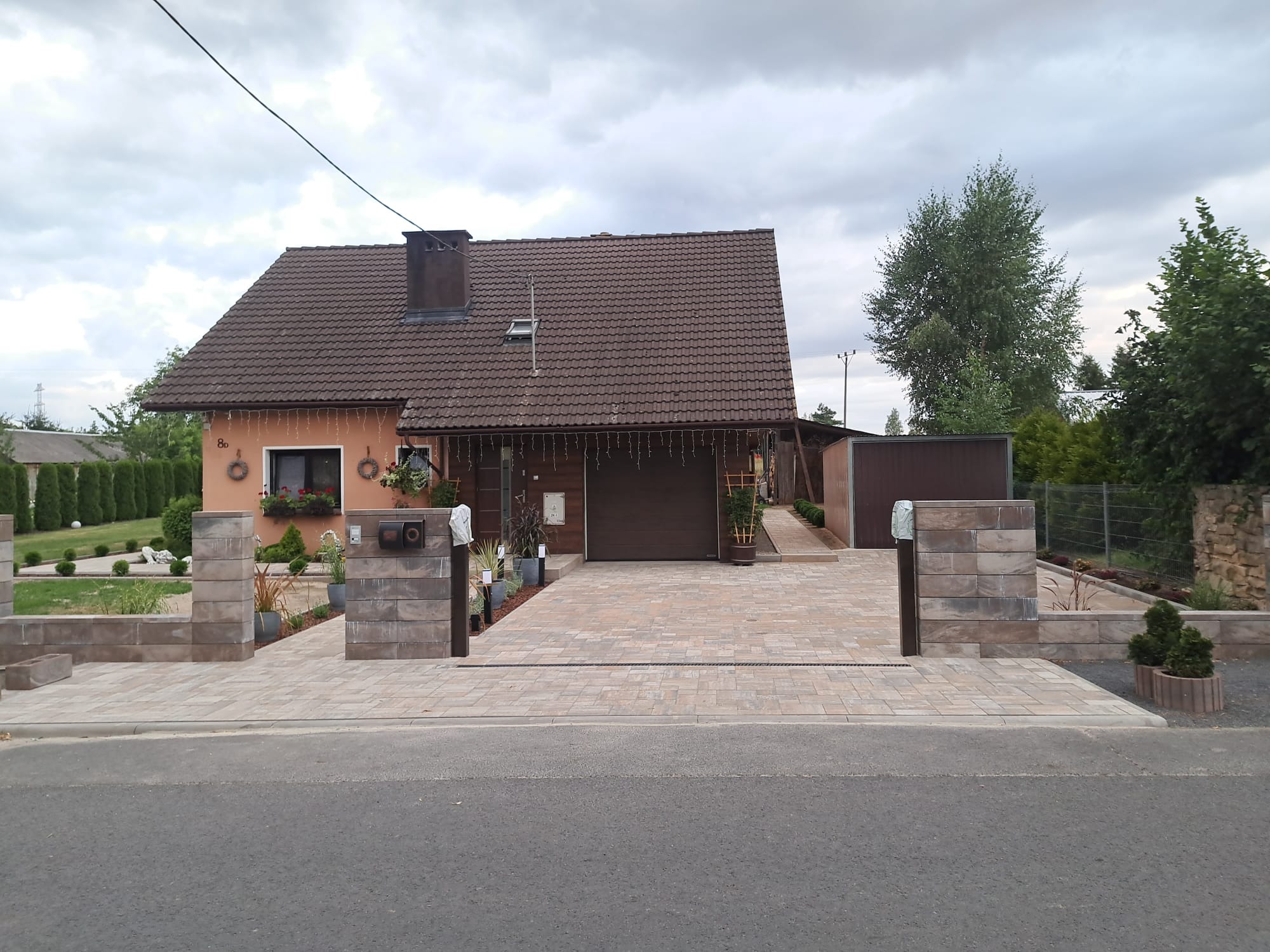 Ułożona kostka brukowa w jasnych odcieniach prowadząca do domu z brązową bramą garażową i zadbanym ogrodem, widoczne słupki z bloków betonowych przy wjeździe.