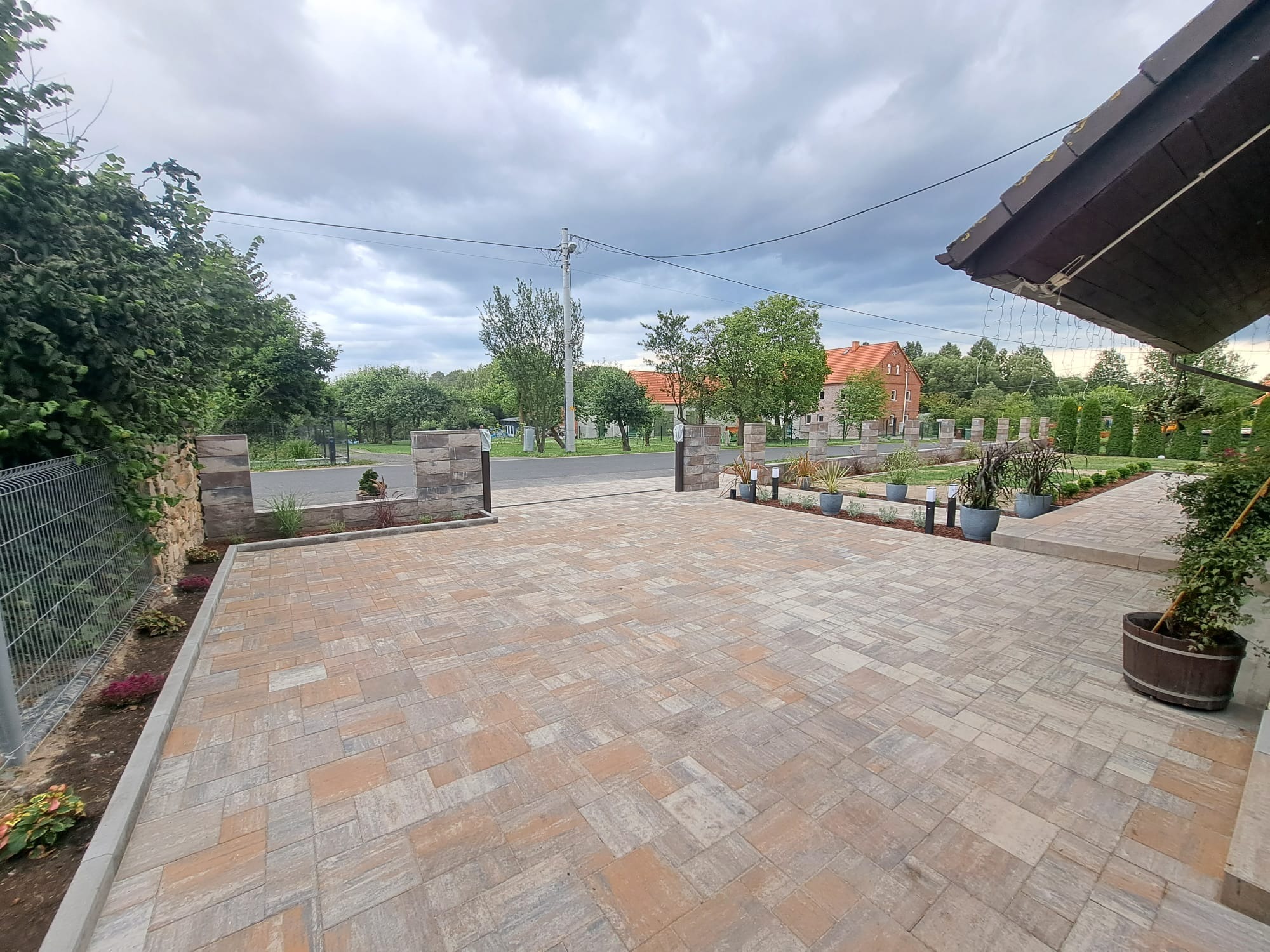 Szeroki podjazd z kostki brukowej w odcieniach szarości i beżu, prowadzący do bramy z kamiennymi słupkami, z widocznymi roślinami rabatowymi po bokach i budynkiem w tle pod pochmurnym niebem.