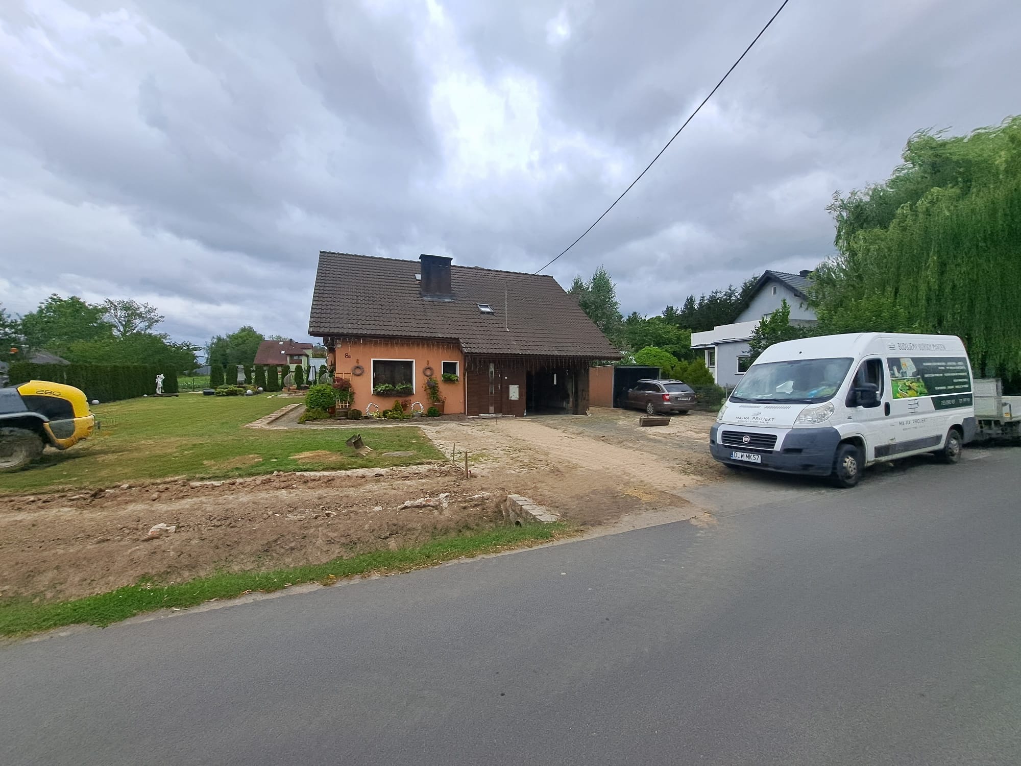 Przygotowanie terenu pod budowę: wyrównana ziemia, ubijarka w tle, budynek mieszkalny i firmowy bus z reklamą na posesji.