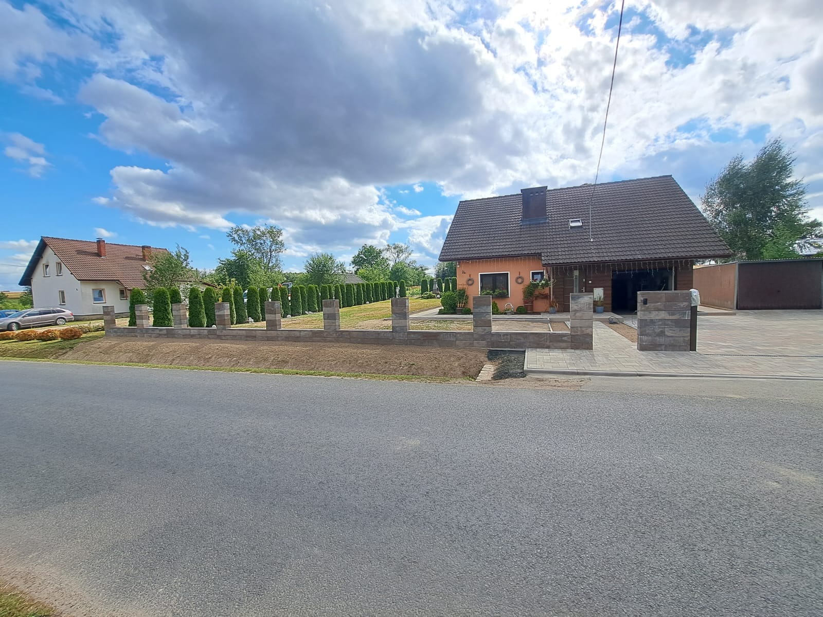 Ułożona kostka brukowa na podjeździe i chodniku prowadzącym do domu z szarej cegły, z niskim murem z identycznej kostki i rzędem tuj w tle, widoczne z poziomu ulicy.