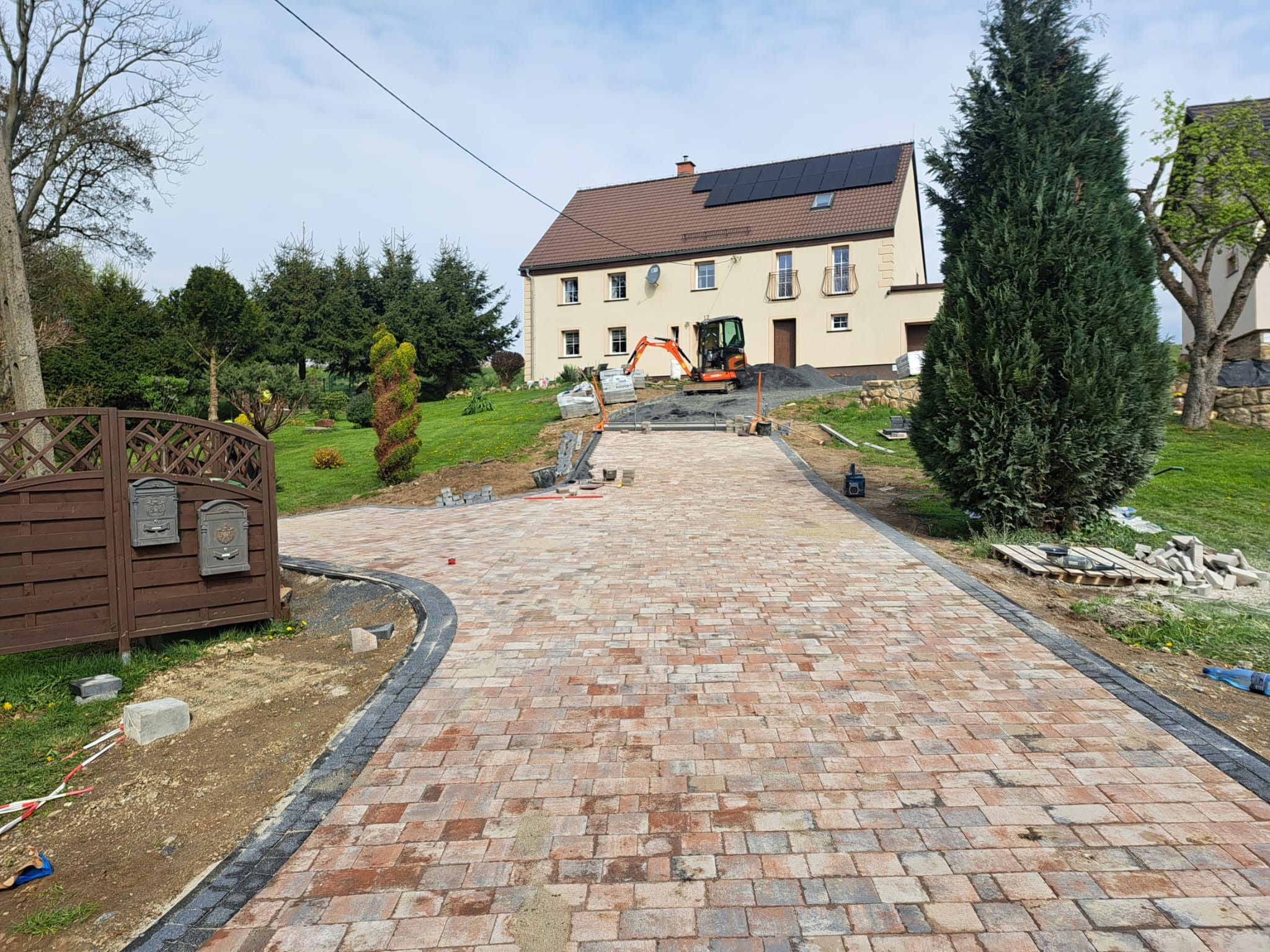 Wjazd do domu z ułożoną kostką w odcieniach czerwieni i beżu, obrzeża wykończone szarą kostką, w tle dom z panelami słonecznymi i minikoparka przygotowująca teren pod dalszą część podjazdu.