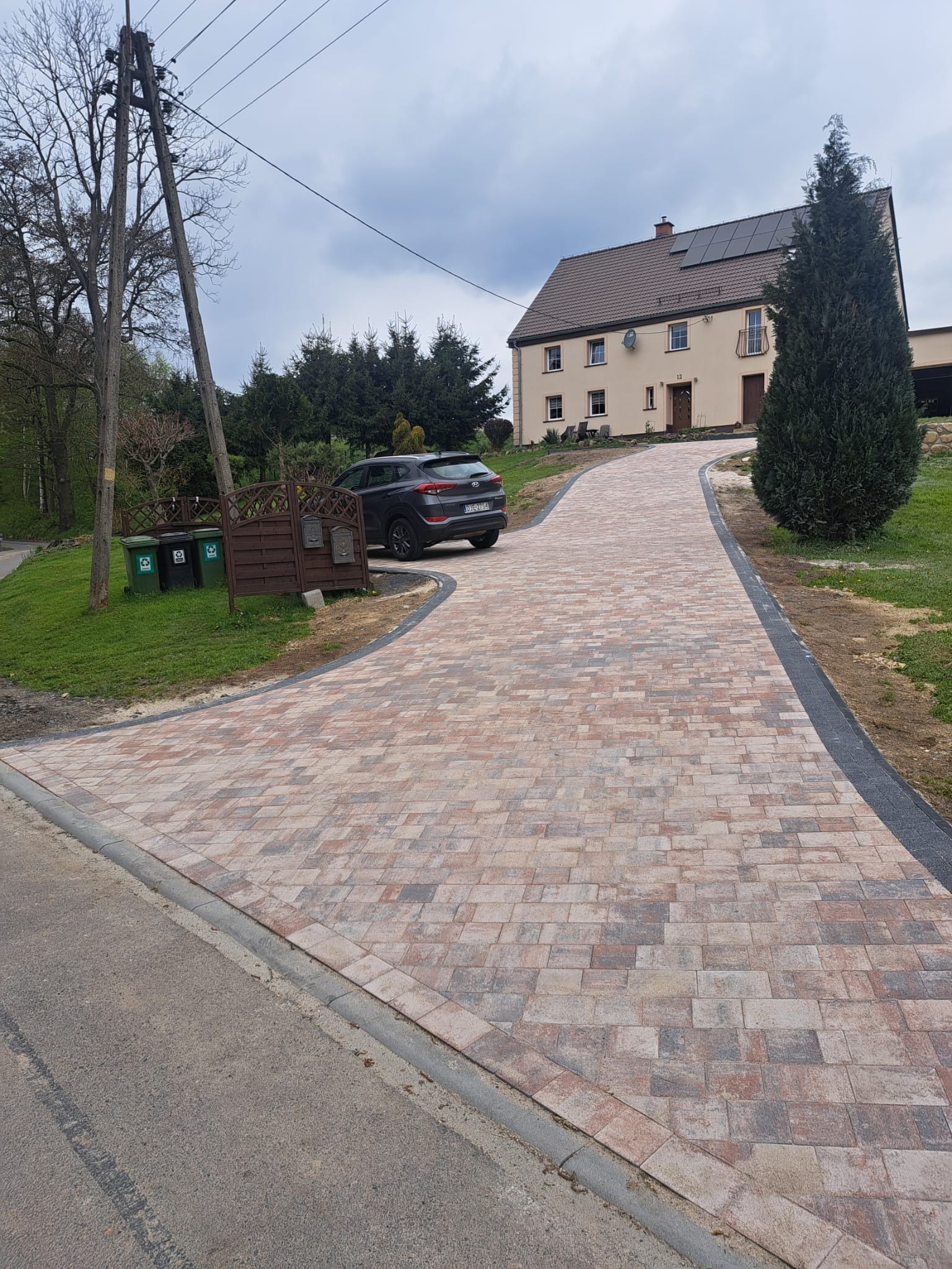 Podjazd z kostki brukowej w odcieniach beżu i czerwieni prowadzący do domu z panelami słonecznymi, widok z poziomu ulicy, szara obwódka z kostki, samochód zaparkowany obok brązowego płotu z trzema...